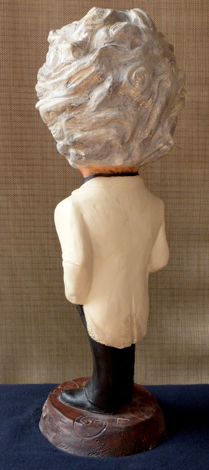 May include: A white ceramic figurine of a person with a white, textured hair style. The figurine is wearing a white jacket and black pants. The figurine is standing on a brown base with a design on it.