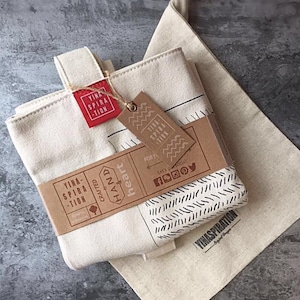 Könnte beinhalten: Cremefarbene Tragetasche mit passendem Kordelzugbeutel. Die Tragetasche hat ein rotes Etikett und ein braunes Papierband mit dem Text "YINA SPIRATION" und "HAND & heart". Das Design umfasst schwarze geometrische Muster und die Worte "100% Baumwolle".