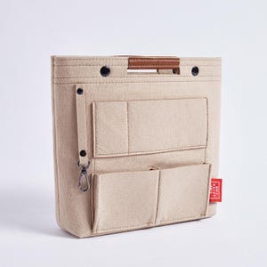 May include: Beige felt bag organizer with multiple pockets and a brown leather handle. Features a small red tag with white text. Includes a metal clasp and black snap closures. Ideal for handbags.