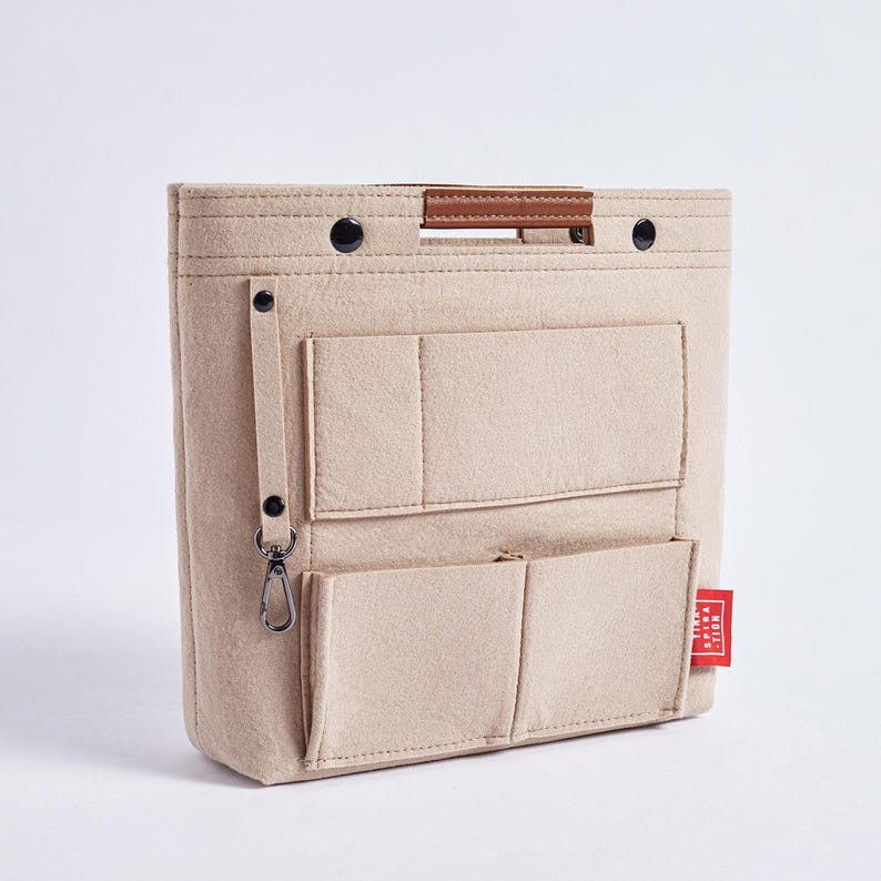 May include: Beige felt bag organizer with multiple pockets and a brown leather handle. Features a small red tag with white text. Includes a metal clasp and black snap closures. Ideal for handbags.