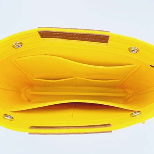 May include: Bright yellow felt bag organizer with multiple pockets and compartments. Features brown leather handles and silver-toned metal rivets. The interior is also yellow, providing a cohesive and organized design.