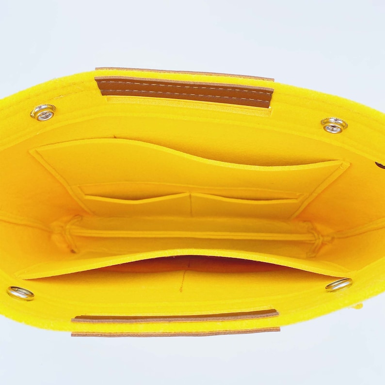 May include: Bright yellow felt bag organizer with multiple pockets and compartments. Features brown leather handles and silver-toned metal rivets. The interior is also yellow, providing a cohesive and organized design.