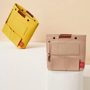 May include: Two felt bag organizers in yellow and beige. The yellow organizer is tilted, showcasing multiple pockets and a brown handle. The beige organizer, upright, features similar pockets, a brown handle, and a small red tag. Both are designed for bag organization.