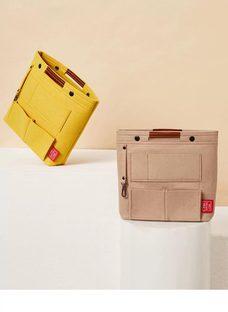 May include: Two felt bag organizers in yellow and beige. The yellow organizer is tilted, showcasing multiple pockets and a brown handle. The beige organizer, upright, features similar pockets, a brown handle, and a small red tag. Both are designed for bag organization.