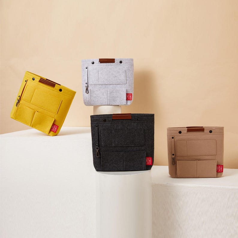 May include: Four felt bag organizers in yellow, gray, black, and tan. Each organizer has a brown handle, multiple pockets, and a small red tag. The organizers are displayed on a white surface against a neutral background.