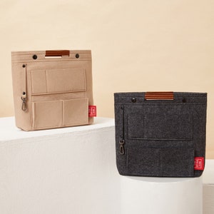 May include: Two felt bag organizers in beige and charcoal gray. Each has multiple pockets, a brown leather handle, and a small red tag with white text. The beige organizer is on the left, and the gray one is on the right.