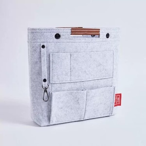 May include: A light gray felt handbag organizer with multiple pockets and a brown leather handle. Features a black carabiner clip and a small red tag. The organizer is designed to keep items neatly arranged.