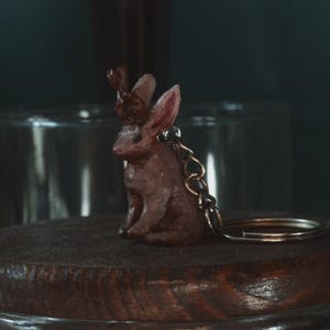 May include: A brown bunny keychain with small antlers, a metal chain, and a key ring. The bunny is sitting on a dark wooden surface. The background is blurred, with a glass jar visible.