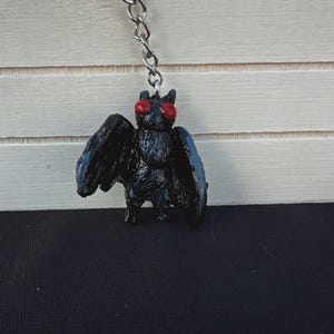 May include: A black keychain featuring a moth-like creature with red eyes and wings. The keychain is attached to a silver chain and ring. The creature has a textured surface and stands on a black surface.