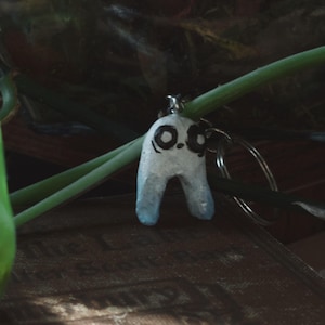 May include: A handmade keychain featuring a white and blue figure with black eyes and a mouth. The keychain is attached to a silver keyring. The figure is hanging in front of a green plant.