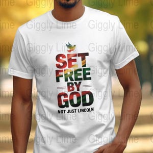 Könnte beinhalten: Weißes T-Shirt mit dem Text "SET FREE BY GOD" in Rot, Gelb, Grün und Schwarz. Der Satz "NOT JUST LINCOLN" steht unter dem Haupttext. Eine kleine Vogelgrafik befindet sich über dem Wort "SET".