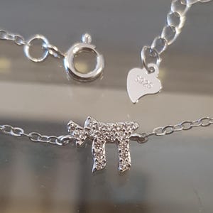 May include: A silver chain necklace with a small pendant shaped like the Hebrew letter 'Chai' with a diamond-like design. The pendant is attached to a silver chain with a heart-shaped tag that says '925'.