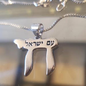 May include: A silver pendant necklace featuring the Hebrew text "Am Yisrael" (People of Israel). The pendant is in the shape of the Hebrew letter "Chai", meaning "life". The necklace has a delicate chain.
