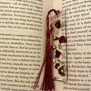 May include: A red and gold bookmark with a tassel. The bookmark is made of clear resin and contains dried flowers and gold leaf. The bookmark is sitting on top of a book with the text "Four Thousand Weeks" and "The Impatient" visible.