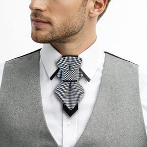 May include: A man in a formal outfit, wearing a gray vest over a white dress shirt and black pants. The outfit includes a black and white patterned ascot tie. The man is seated in a black chair.