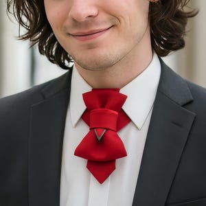 May include: A dark charcoal suit jacket worn over a crisp white button-down shirt, featuring a prominent, structured red fabric neck accessory tied in a layered bow shape with a small metallic V-shaped accent at the centre of the knot.