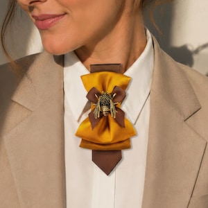 May include: A mustard yellow and brown bow tie with a gold bee embellishment. The bow tie is worn over a white collared shirt and a tan blazer. The bee has intricate detailing.