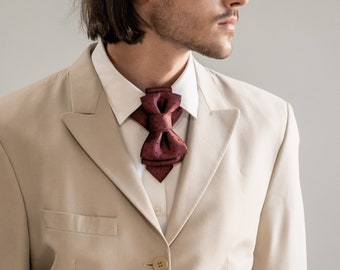 Peut inclure: Une personne portant une veste de costume beige avec un nœud papillon bordeaux. La veste a deux boutons dorés et une ceinture marron.