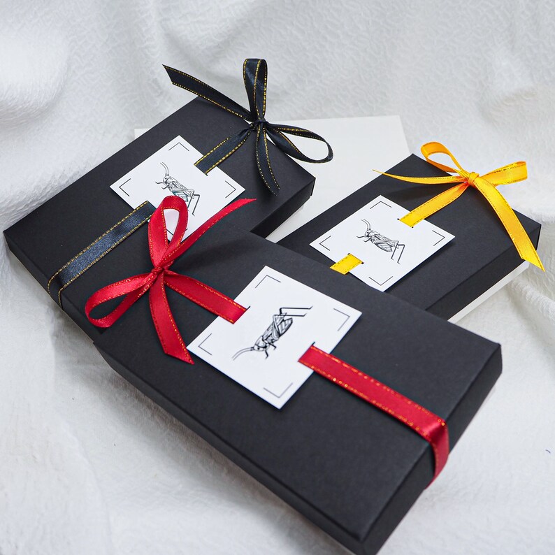 May include: Three black gift boxes with red, yellow, and black ribbon. Each box has a white label with a black and white illustration of a cricket.