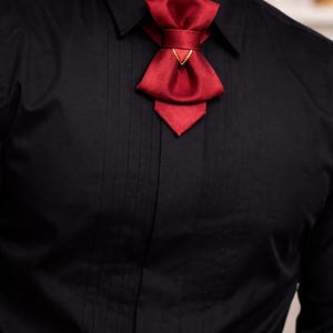 May include: A solid black button-down shirt with vertical pleats along the front placket, featuring a deep red satin bow tie accessory secured at the collar with a small gold-toned triangular metal accent piece centred on the knot.
