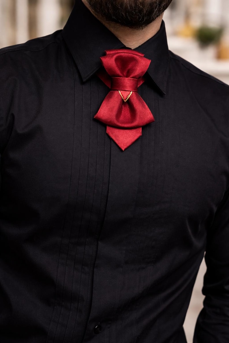 May include: A solid black button-down shirt with vertical pleats along the front placket, featuring a deep red satin bow tie accessory secured at the collar with a small gold-toned triangular metal accent piece centred on the knot.