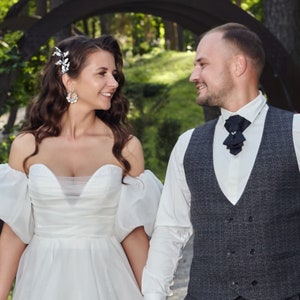 May include: A bride and groom are smiling at each other. The bride wears a white off-the-shoulder wedding dress with puffy sleeves and a jeweled hairpiece. The groom wears a gray vest over a white shirt and a black ascot.