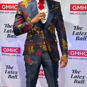 May include: A person wearing a wide-brimmed red hat with a feather, round sunglasses, a white shirt with a red bowtie, and a dark blue blazer featuring a floral pattern in red and gold. The outfit includes dark blue trousers and black dress shoes.