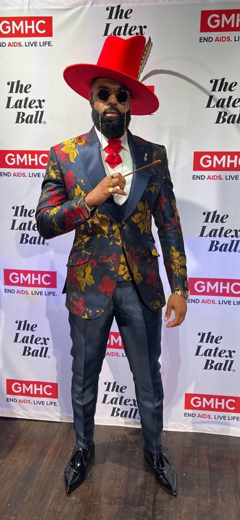 May include: A person wearing a wide-brimmed red hat with a feather, round sunglasses, a white shirt with a red bowtie, and a dark blue blazer featuring a floral pattern in red and gold. The outfit includes dark blue trousers and black dress shoes.
