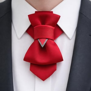 May include: A bright red satin cravat featuring a structured bow design with a central horizontal band and a downward-pointing triangular silver-toned metal accent piece with a ribbed texture, worn over a crisp white collared shirt and dark blazer.