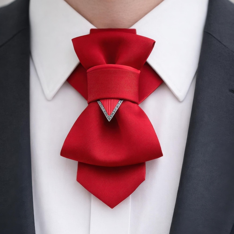 May include: A bright red satin cravat featuring a structured bow design with a central horizontal band and a downward-pointing triangular silver-toned metal accent piece with a ribbed texture, worn over a crisp white collared shirt and dark blazer.