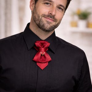 May include: A deep red satin cravat featuring a layered bow design with a central gold-toned triangular accent, worn over a black button-down shirt with a pointed collar and vertical pleats along the front placket, creating a formal appearance.