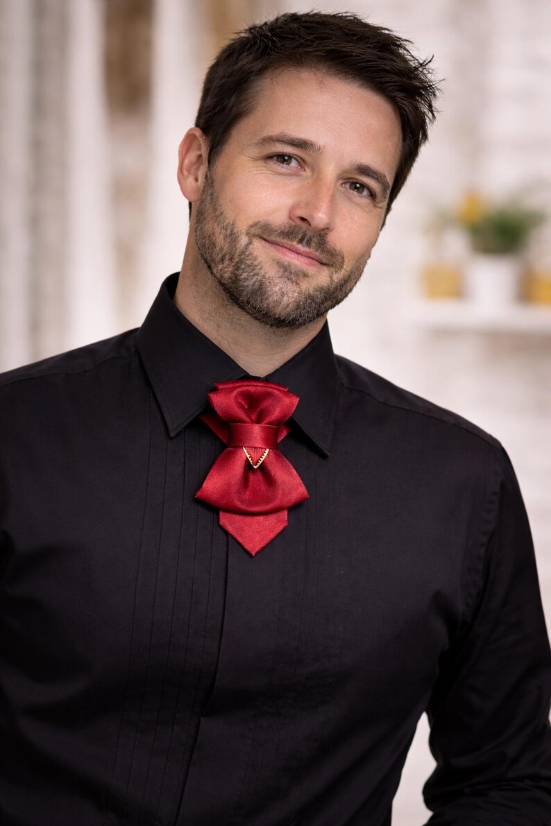 May include: A deep red satin cravat featuring a layered bow design with a central gold-toned triangular accent, worn over a black button-down shirt with a pointed collar and vertical pleats along the front placket, creating a formal appearance.