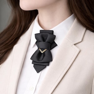 May include: A black bow tie with a gold-colored accent, worn over a white collared shirt and a light beige blazer. The bow tie has a layered design and a geometric gold detail.