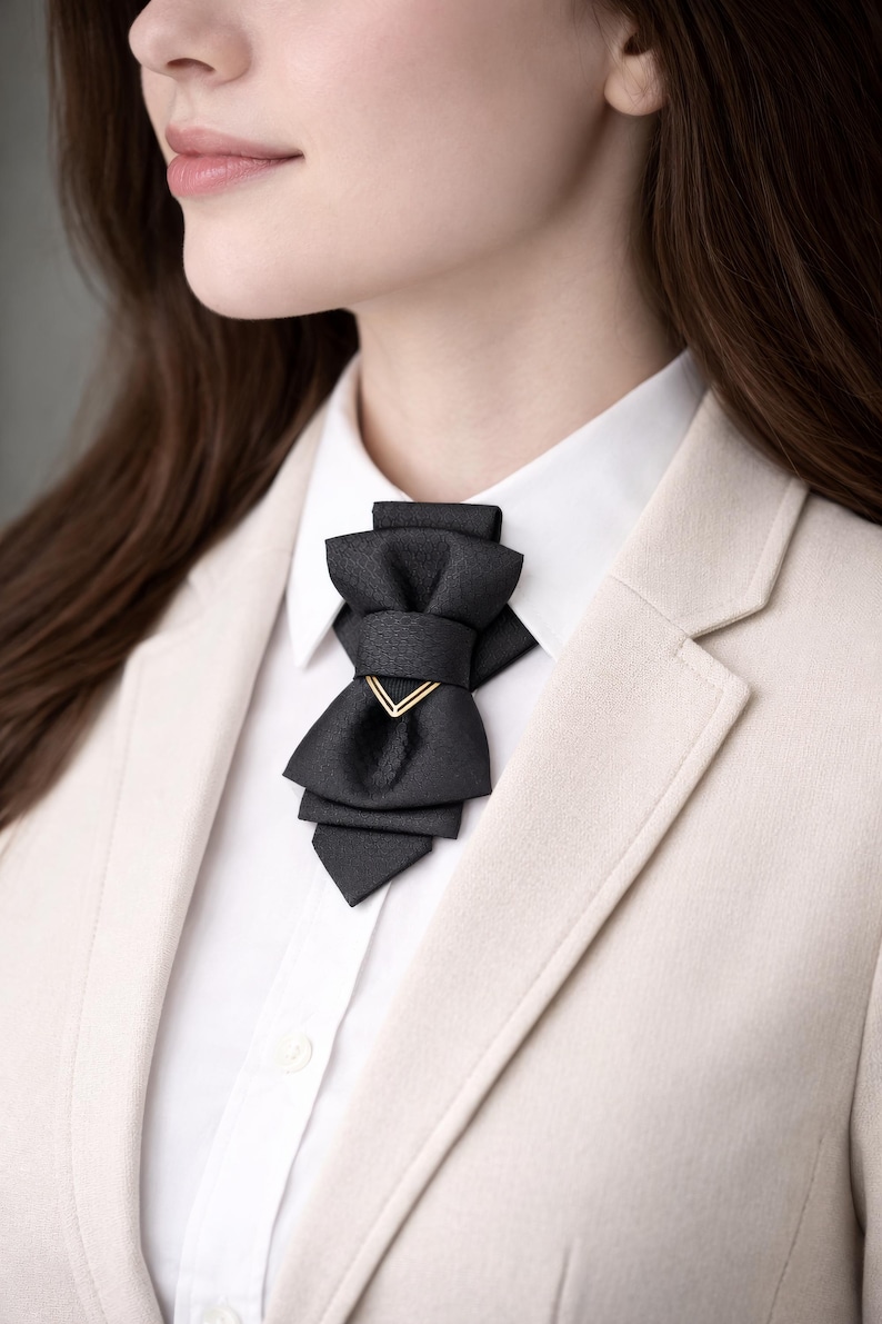May include: A black bow tie with a gold-colored accent, worn over a white collared shirt and a light beige blazer. The bow tie has a layered design and a geometric gold detail.