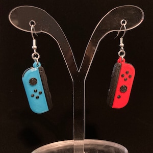 May include: A pair of earrings featuring a blue and black Nintendo Switch Joy-Con controller on the left and a red and black Nintendo Switch Joy-Con controller on the right.