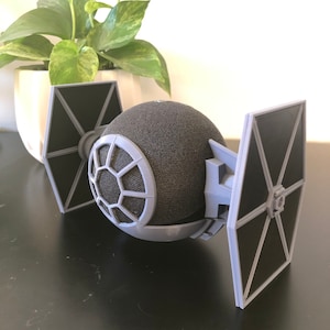 May include: A gray 3D printed TIE fighter stand for a smart speaker. The stand has two wings and a round opening in the center for the speaker.