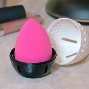 May include: A pink makeup sponge in a black and white drying holder. The holder has a mesh design to allow air circulation.
