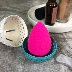 May include: A pink makeup sponge sits inside a teal blue and white plastic holder with a circular design. The holder has a white base and a teal blue top with a circular pattern.