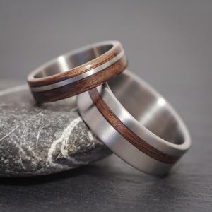 Könnte beinhalten: Zwei Eheringe mit einem silbernen Band und einer dunklen Holzeinlage. Ein Ring ist auf dem anderen gestapelt und liegt auf einem grauen Stein. Die Ringe haben eine gebürstete Metalloberfläche und ein minimalistisches Design.