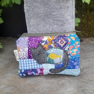 May include: A small patchwork fabric zippered pouch with a grey felt cat appliqué with a yellow fish inside a blue and green circle. The pouch is made of various colourful fabrics with floral, geometric, and polka dot patterns.
