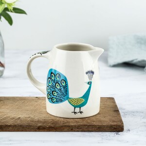 May include: A white ceramic pitcher with a peacock design. The peacock is blue, green, and yellow with a black outline. The pitcher has a handle and a spout.