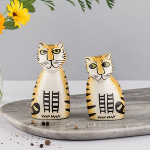 Könnte beinhalten: Ein Set aus zwei Keramik-Salz- und Pfefferstreuern in Tigerform. Die Streuer sind mit orangefarbenen und schwarzen Streifen bemalt und haben schwarze Augen und Schnurrhaare. Sie stehen auf einer grauen Marmorfläche.