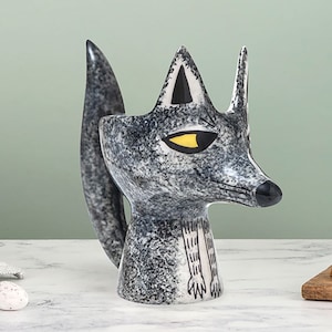 Puede incluir: Una taza de cerámica con forma de zorro con un esmalte moteado en blanco y negro. El zorro tiene un ojo amarillo y una nariz negra. La taza es una gran adición a cualquier cocina.