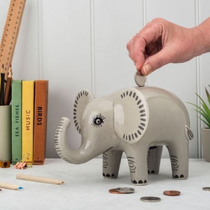 May include: A grey ceramic elephant money box with black and white details. A hand is placing a coin in the slot on top of the elephant's head.