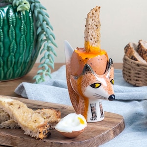 Peut inclure: Un porte-œuf en céramique en forme de renard avec un motif de renard brun et orange. Le porte-œuf contient un œuf dur avec un jaune d'œuf coulant. Un morceau de pain grillé dépasse du haut du porte-œuf.