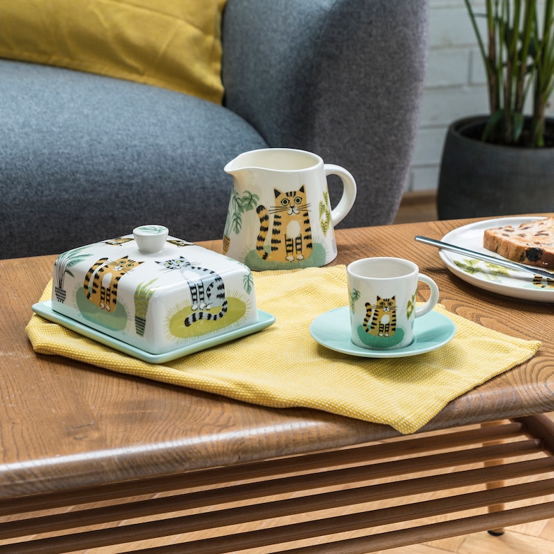 Puede incluir: Un juego de vajilla de cocina de cer&aacute;mica con un dise&ntilde;o de gato caprichoso. El juego incluye una mantequillera, una jarra de crema y una taza de t&eacute; y su platillo. Las ilustraciones de gatos son en tonos de naranja, negro y blanco, con acentos verdes. El fondo es azul claro.