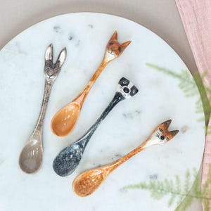 May include: Four decorative spoons with animal-shaped handles. The spoons feature a bunny, two foxes, and a panda bear. The spoons are on a white marble surface. The spoons have brown, black, and white details.