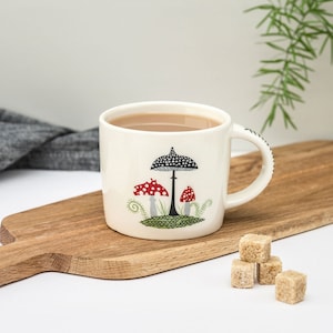 May include: White ceramic mug with a black and white mushroom design and red and white mushrooms. The mug is filled with tea and is sitting on a wooden cutting board with sugar cubes.