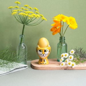 May include: Yellow ceramic egg cup shaped like a cat with black eyes and a black nose. The egg cup is holding a white egg. The egg cup is on a wooden cutting board with a vase of yellow flowers and a vase of white flowers in the background.