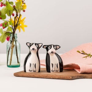 May include: A pair of ceramic salt and pepper shakers shaped like black and white dogs. The shakers have a glossy finish and are sitting on a wooden cutting board. A vase of yellow flowers and a pink cloth are in the background.